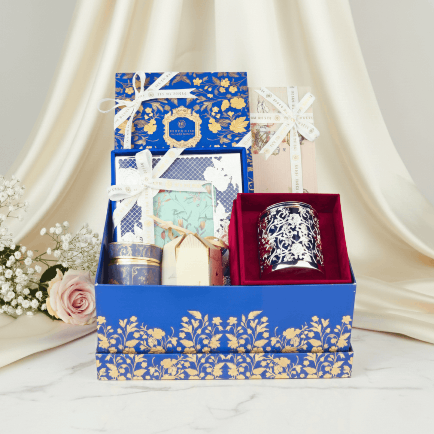 Premium Fleur de Lis gift hamper styled with soft draped fabric, showcasing luxury candle, gourmet confections, and handcrafted gift boxes in an elegant presentation.